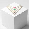 Four gold-tone butterfly necklaces with colorful gemstone wings are arranged on a white display cube. The necklaces feature pink, green, silver, and purple butterfly pendants.