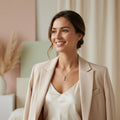 A smiling woman in a cream satin top and beige blazer wears a delicate gold necklace with a bar pendant. The image is a promotional ad with text reading 'Elevate your everyday with a touch of timeless brilliance.