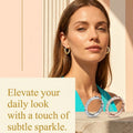 A promotional fashion graphic featuring a woman with long light brown hair wearing small gold hoop earrings and a turquoise top, with a pair of round silver-framed sunglasses resting on a stone ledge. Text on the left reads, 'Elevate your daily look with a touch of subtle sparkle.