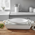 A set of white ceramic baking dishes with black rim detailing, stacked on a rustic wooden table next to a sprig of rosemary and a beige cloth, with a softly blurred kitchen background.