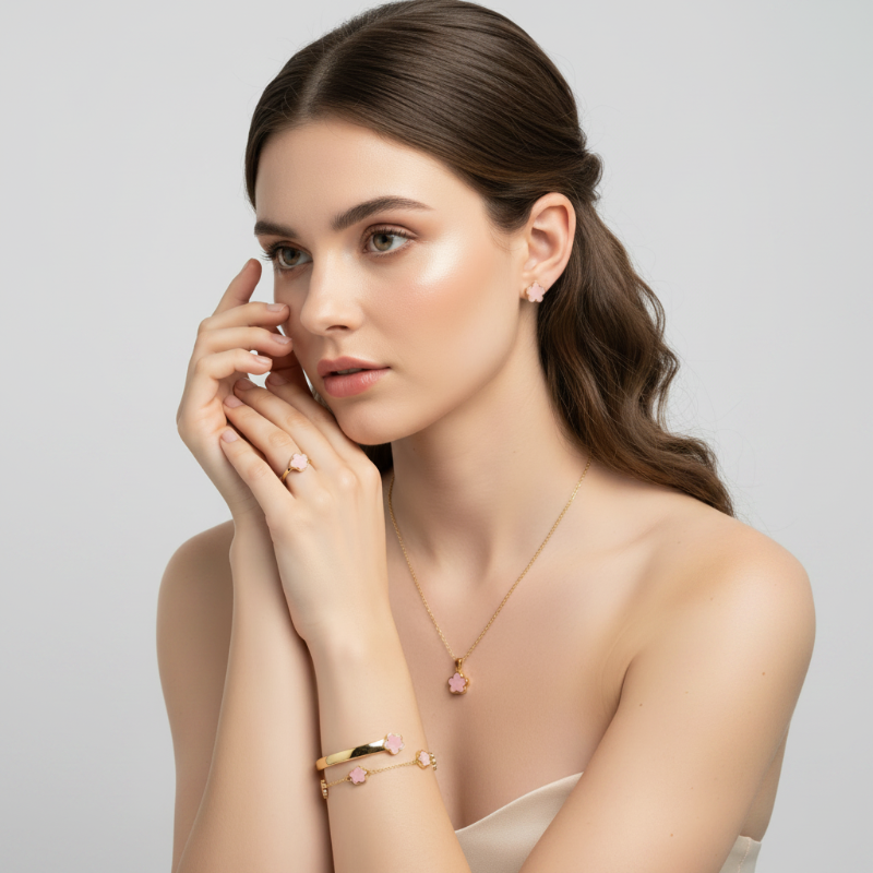 A woman with brown hair models a gold jewelry set featuring a delicate necklace with a pink flower pendant, a matching bracelet, and a ring with a pink gemstone, against a clean white background.