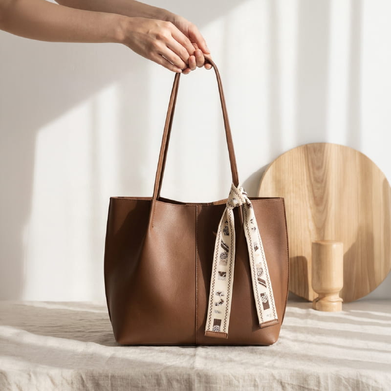 A brown leather tote bag with long handles and a cream patterned scarf tied around one handle, held by a person's hands in a softly lit room with a wooden board and vase in the background.