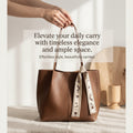 A brown leather tote bag with double top handles and a patterned cream ribbon tied around one handle, held by a person's hands in a softly lit interior setting. Text overlay reads: 'Elevate your daily carry with timeless elegance and ample space.